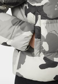 Pildīta jaka pelēkā un baltā abstraktā rakstā; aprīkota ar rāvējslēdzēja kabatu un elastīgiem piedurknēm, lai nodrošinātu piemērotību un siltumu. Strukturēta auduma.