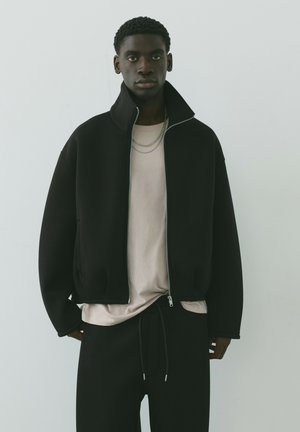Jeune homme portant une veste zippée noire sur une chemise beige clair et un pantalon noir, debout devant un fond uni gris clair.