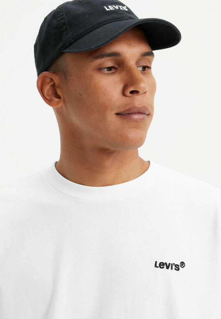 Giovane uomo con cappellino nero Levi's e t-shirt bianca Levi's, che guarda leggermente a sinistra su uno sfondo semplice.