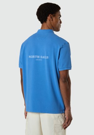 Hombre de espaldas con una camiseta tipo polo azul brillante con el texto "North Sails Archive" en la parte trasera y pantalones cortos cargo beige claro.