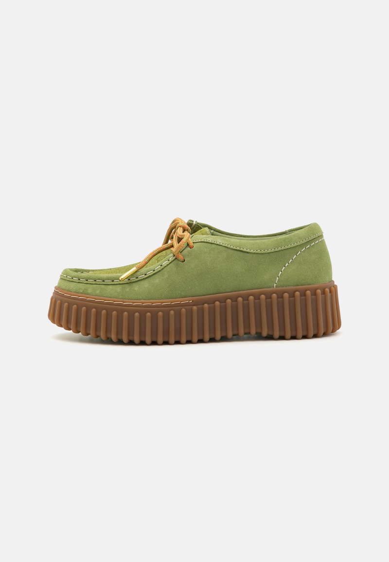 Zapato de ante verde con puntera redonda, suela marrón estriada y cordones naranjas. Presenta costuras en contraste a lo largo de los bordes.