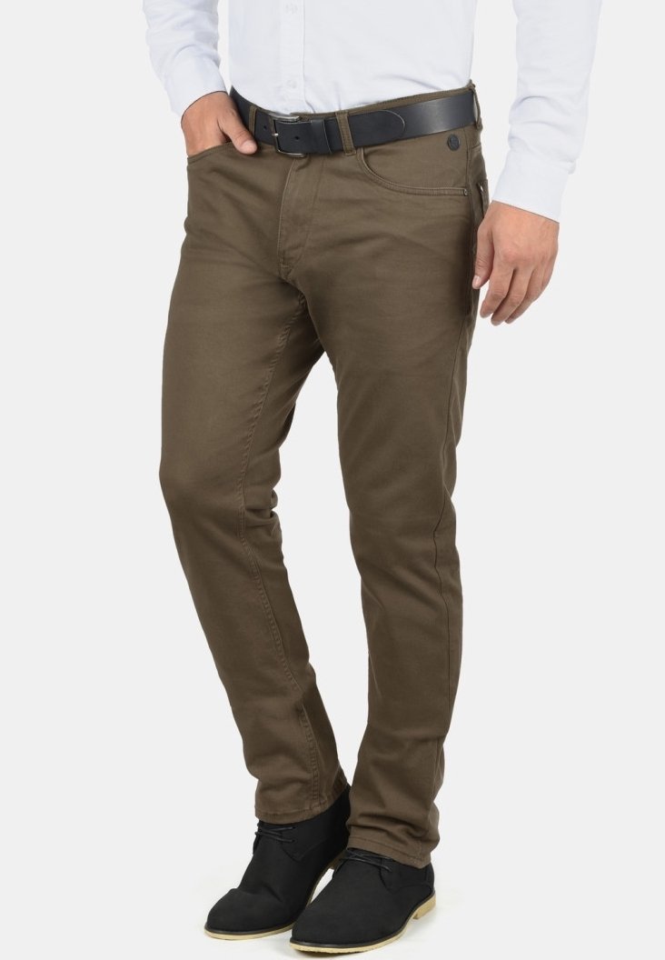 Homme portant un pantalon slim marron, une ceinture noire, des chaussures noires et une chemise blanche à manches longues, une main dans la poche.