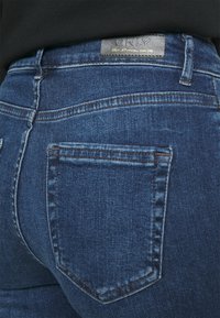 Närbild av bakfickan och midjebandet på blå denimjeans med en svart läderlapp märkt "ONLY" på en person som bär en svart topp.