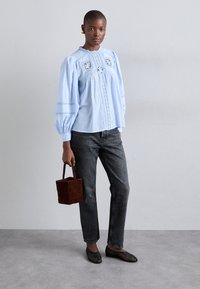 Nainen, jolla on yllään sininen pitsiyksityiskohdalla varustettu paita, mustat farkut ja mustat kudotut ballerinat, pitelee pientä ruskeaa neliömäistä käsilaukkua seisten yksinkertaista harmaata seinää vasten.