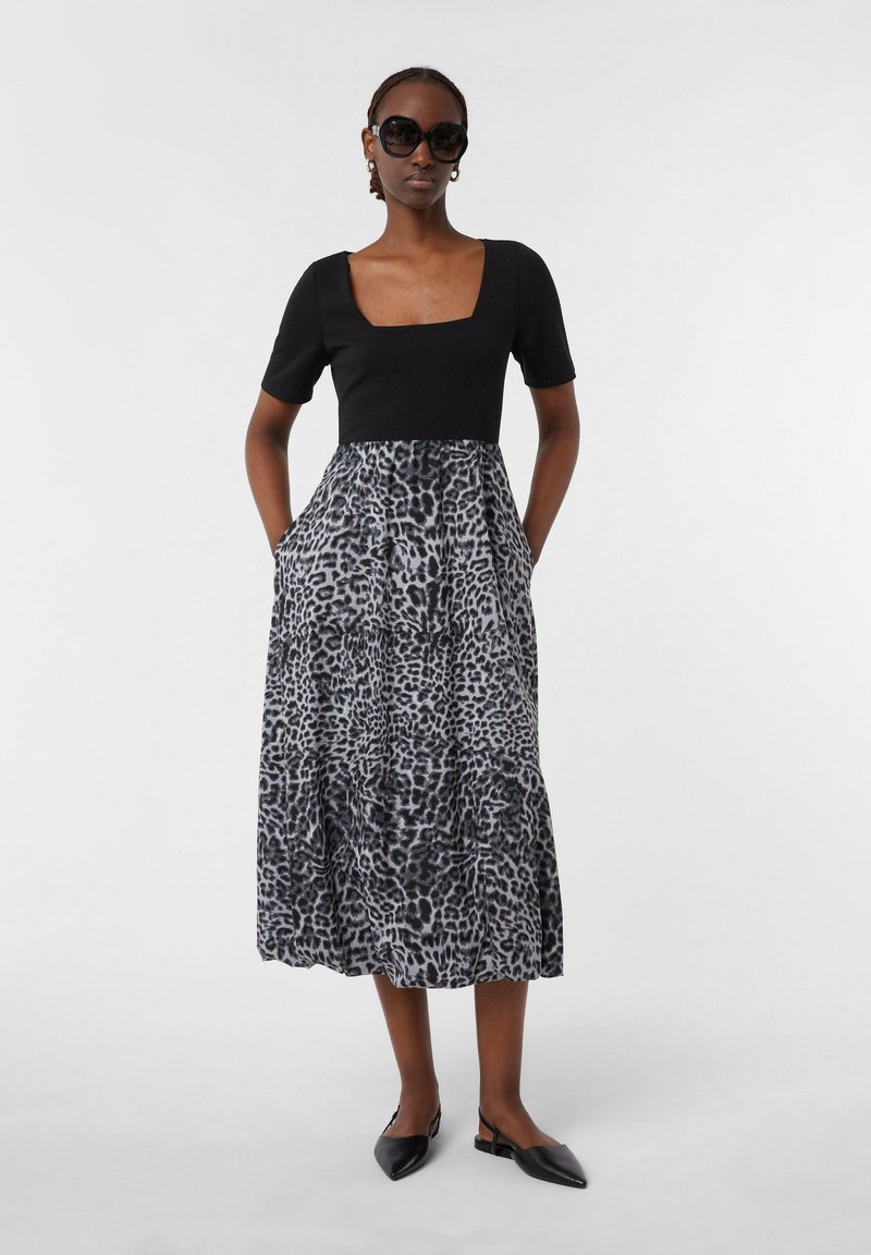 Schwarzes Kurzarmkleid mit quadratischem Ausschnitt, Mieder mit grauem Leopardenmuster, Taschen und flachen schwarzen Schuhen.