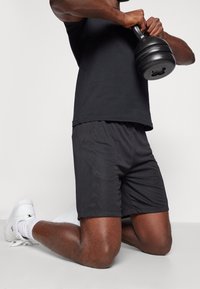 Černé sportovní šortky s hladkou texturou a jemným vzorem loga na boku, spárované s černým tričkem a bílými sportovními botami.