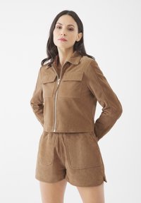 Giubbino corto in suede marrone con due tasche sul petto e zip argentata. Abbinato a pantaloni corti in suede coordinati con tasche frontali e spacchi laterali.