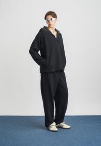 Moletom preto de fleece com fecho no pescoço, emparelhado com calças combinando. Apresenta uma superfície texturizada, bolsos laterais e ténis brancos clássicos.