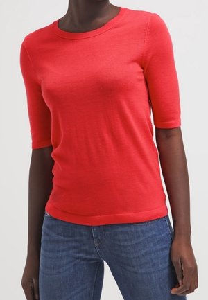Person trägt einen eng anliegenden roten Kurzarm-Pullover und blaue Jeans, steht vor hellem, einfarbigem Hintergrund, Oberkörper zu sehen.