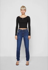 Jovem com cabelo curto loiro, usando uma blusa preta de manga longa cropped, jeans azuis e sandálias pretas com tiras no tornozelo, em pé.