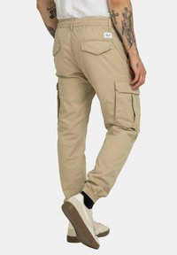 Beige Cargo-Hosen mit elastischen Bündchen, seitlichen Taschen und einem Patch auf der Rückseite. Glattes Material mit entspanntem Schnitt und schmal zulaufendem Bein.