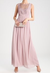 Dlouhá růžová večerní šaty s plisovaným korzetem a průhledným, zdobeným výstřihem. Modelka drží stříbrnou kabelku a má na sobě nahé lodičky.