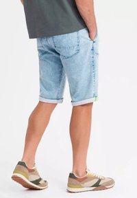 Pantalones cortos de mezclilla azul claro con puños doblados, que presentan dos bolsillos traseros y un acento verde neón. Combinados con zapatillas deportivas en beige y verde.