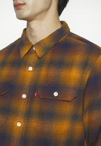 Gros plan d'une personne portant une chemise à carreaux jaune et bleu marine avec des boutons blancs et deux poches poitrine.