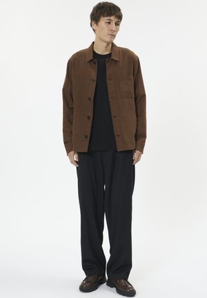 Barna gombos ing egyetlen mellzsebbel, fekete póló fölött viselve, fekete nadrággal és barna cipővel párosítva.