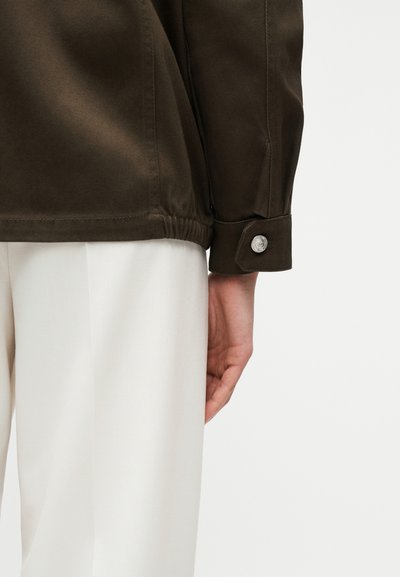 Κοντινό πλάνο του χεριού ενός ατόμου που ξεκουράζεται δίπλα στο πλευρό του, φοράει σκούρο καφέ πουκάμισο με μακριά μανίκια και μανσέτα με κουμπί, καθώς και λευκό παντελόνι.