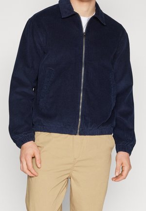 Mężczyzna w granatowej sztruksowej kurtce na zamek i beżowych spodniach, stojący na jednolitym tle.