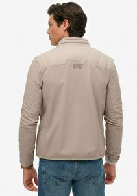 Veste beige clair avec col montant, panneaux supérieurs matelassés et logo "SUPER DRY" brodé au dos. Comprend des poches latérales.