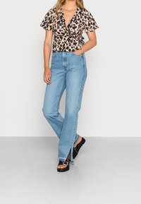 Femme portant un haut enveloppant à motif léopard, un jean taille haute bleu clair avec des fentes sur les côtés, et des sandales noires avec des détails de boucle.