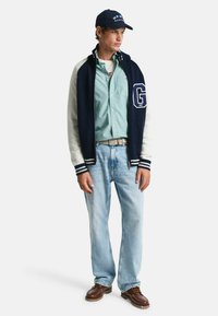 Marineblauwe en witte gebreide varsityjack met een groot 'G' logo, lichtgroen shirt, lichtblauwe spijkerbroek, bruine schoenen en een donkere pet.