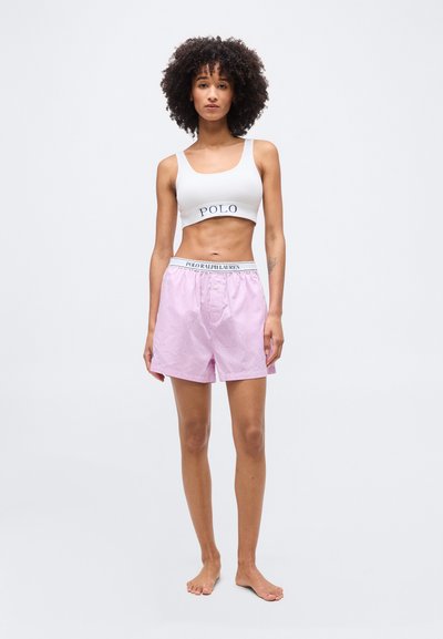 Γυναίκα με σγουρά μαλλιά που φοράει λευκό cropped top με τη λέξη «POLO» και ροζ ριγέ σορτς με λάστιχο μέσης «POLO RALPH LAUREN», όρθια και ξυπόλητη.