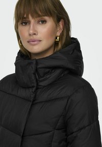 Doudoune noire avec un col haut, présentant des sections matelassées horizontales et une texture lisse. Détail du visage du modèle et de grandes boucles d'oreilles.