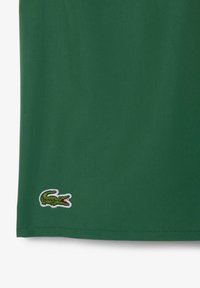 Tissu en coton vert avec une texture lisse ; présente un logo de crocodile brodé blanc près de l'ourlet. Design simple sans motifs supplémentaires.