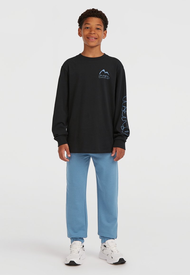 Smiling boy stands wearing black long-sleeve shirt with blue logo, light blue joggers, and white sneakers against a plain white background.