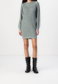 Femme portant une mini-robe tricotée gris clair à manches longues et des bottes en cuir noires mi-mollet avec des boucles, debout devant un fond blanc uni.