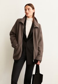 Femme portant une veste en polaire marron à fermeture éclair par-dessus un blazer noir et un turtleneck blanc, tenant un sac fourre-tout noir, debout contre un mur uni.