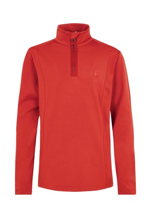 Jong persoon staat, draagt een rood shirt met lange mouwen en een kwartrits, zwarte broek, zwarte laarzen en een zwarte muts.