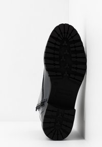 Czarna podeszwa buta z głębokim bieżnikiem i widocznym zamkiem błyskawicznym po boku, ustawiona pionowo na tle białego narożnika.