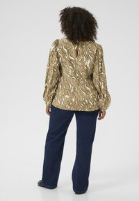 Halenka s dlouhým rukávem v hořčicové barvě s abstraktním bílým a černým vzorem, se zadním výstřihem ve tvaru klíčku a nabíranými rukávy, ve spojení s tmavými džíny.