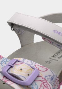 Κοντινή λήψη λουριού άσπρου σανδαλιού με μοβ πόρπη και πολύχρωμα σχέδια κινουμένων σχεδίων στο λουρί.