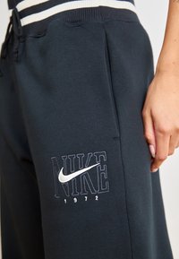 Černé bavlněné tepláky Nike s bílým vyšitým logem, s akcentem z roku 1972 a bočními kapsami. Pas je zdoben bílými pruhy.