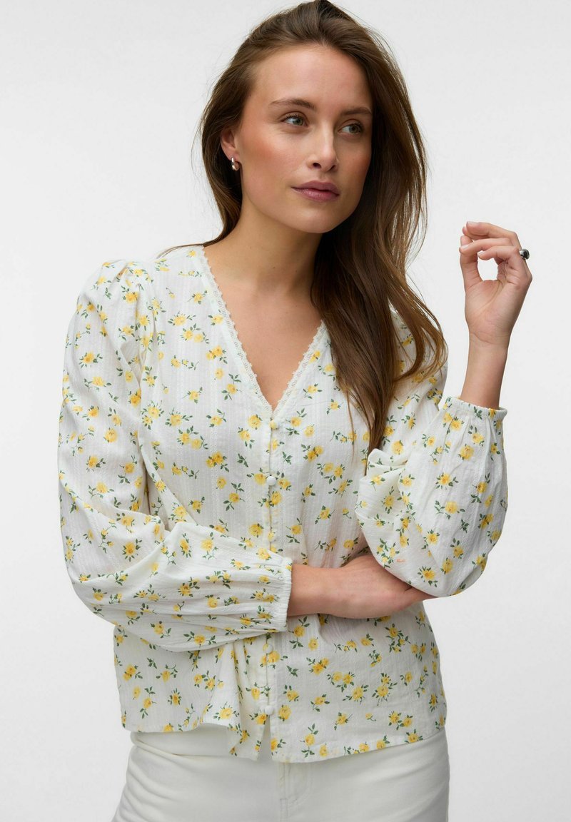 Femme aux longs cheveux bruns portant un chemisier blanc à imprimé floral jaune et manches longues bouffantes, posant devant un fond uni.