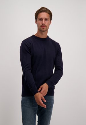 Marineblauwe gebreide trui met een ronde hals. Lange mouwen, ribgebreide manchetten en onderrand. Gecombineerd met lichtblauwe spijkerbroek. Eenvoudig, klassiek ontwerp.