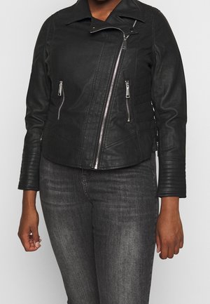 Femme portant une veste en cuir noire asymétrique zippée avec poches zippées et détails matelassés aux manches, assortie à un jean gris foncé.
