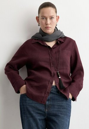 Mujer con cabello peinado hacia atrás lleva una camisa de botones color rojo oscuro, jeans azules, bufanda gris alrededor del cuello, collar largo de cuentas y pendientes de aro plateados.