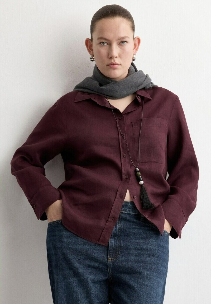 Femme aux cheveux tirés en arrière porte une chemise boutonnée rouge foncé, un jean bleu, une écharpe grise autour du cou, un long collier de perles et des boucles d'oreilles argentées en forme d'anneau.