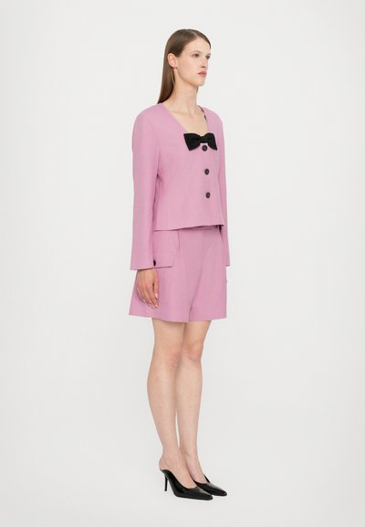 Pink cropped blazer with black bow tie detail, button closures, and matching pink shorts. Model wears black high-heeled shoes.