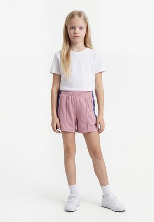 Blond pige står, iført hvid T-shirt, pink atletikshorts med sidelister, hvide sokker og hvide sneakers på en ensfarvet baggrund.