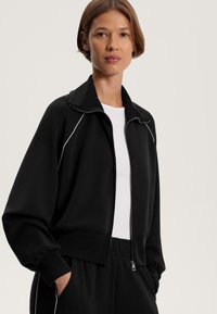 Fekete cipzáros dzseki magas gallérral és fehér díszítéssel. Puha anyagú, laza fazonú, gumis ujjaival, alatta fehér pólóval.