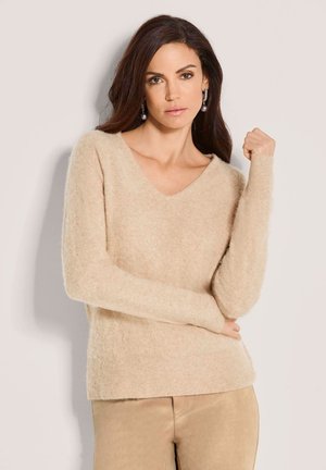 Vrouw met lang donker haar, gekleed in een beige pluizige trui en bijpassende broek, poserend met één arm gebogen bij de elleboog tegen een effen lichte achtergrond.