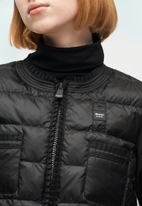 Κοντά κόκκινα μαλλιά, μαύρο καπιτονέ σακάκι με μπροστινές τσέπες και ορατό φερμουάρ, φοράει μαύρο ζιβάγκο από κάτω.