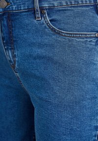 Jeans de denim azul con un acabado texturizado, que cuentan con un bolsillo frontal, botón de metal y detalle de costura en contraste.