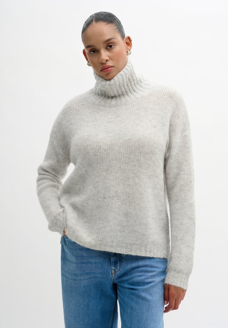 Cream turtleneck sweater made of soft knit fabric, featuring ribbed texture and long sleeves, paired with light blue denim jeans.