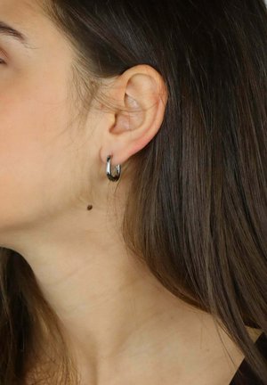 Orecchino a cerchio in argento con una finitura texturizzata e un design curvo, indossato su un modello. Capelli castani leggermente mossi, con una neo visibile sul collo.