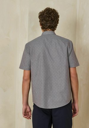 Man with curly hair wearing a short-sleeve gray shirt with a subtle dotted pattern, standing facing a beige wall.