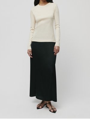 Nainen, joka käyttää kermaisen väristä pitkähihaista paitaa, mustaa nilkkapituista hametta ja mustia remmikypsyisiä litteitä sandaaleja, seisoo yksinkertaisen harmaan taustan edessä.